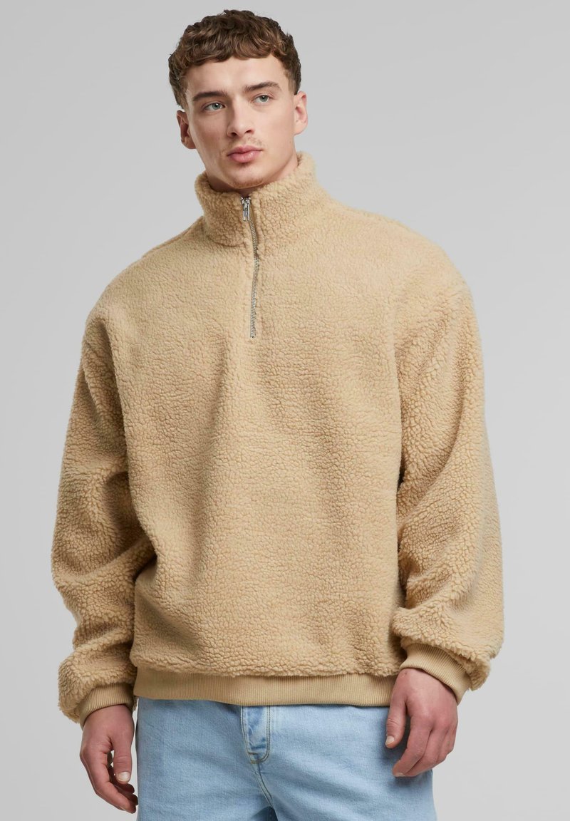 Beige fleece sweater med høj krave og kvart-lynlås, struktureret overflade og elastiske manchetter, båret med lyseblå jeans.
