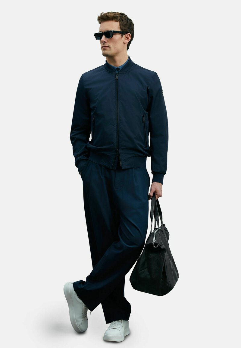 Marineblauer Bomberjacke mit aufstehendem Kragen, lässigen Hosen, weißen Sneakers und einer schwarzen Tasche. Urbanes Design mit minimalistischen Akzenten.