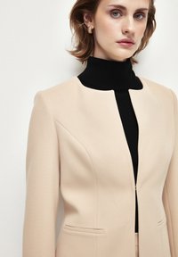 Blazer beige strutturato con una texture liscia, dotato di chiusura con zip, due tasche frontali e un collo alto, indossato sopra un dolcevita nero.