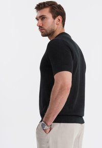 Suéter negro de punto de manga corta con ribete en el bajo, corte relajado y un patrón texturizado, combinado con pantalones de color claro y un reloj.