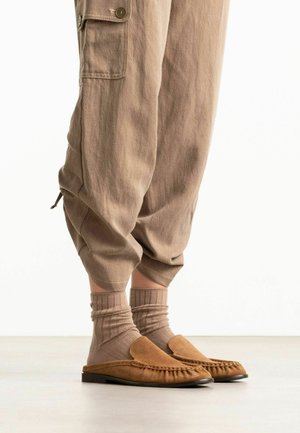 Lower legs wearing beige cuffed pants, ribbed mid-calf socks, and tan suede moccasin slippers on a light floor.