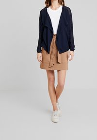 Granatowy kardigan z otwartym przodem i teksturowanymi rękawami, zestawiony z beżową spódnicą do kolan z wiązanym paskiem. Całość dopełniają białe sneakersy.