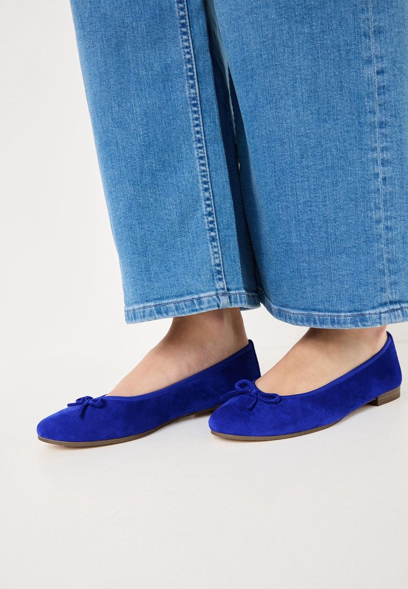 Ballerines en daim bleu avec petits nœuds, portées avec un jean large en denim bleu clair.