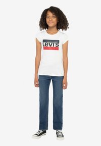 Baltas trumpomis rankovėmis marškinėlis su juodu ir raudonu „Levi's“ logotipu, derinamas su mėlynais džinsais ir juodais sportbačiais.