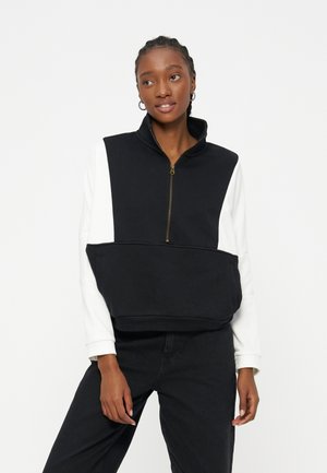 Νεαρή γυναίκα με πλεξούδες φοράει ασπρόμαυρο φούτερ με φερμουάρ και μεγάλη τσέπη μπροστά, στέκεται μπροστά σε απλό λευκό φόντο.