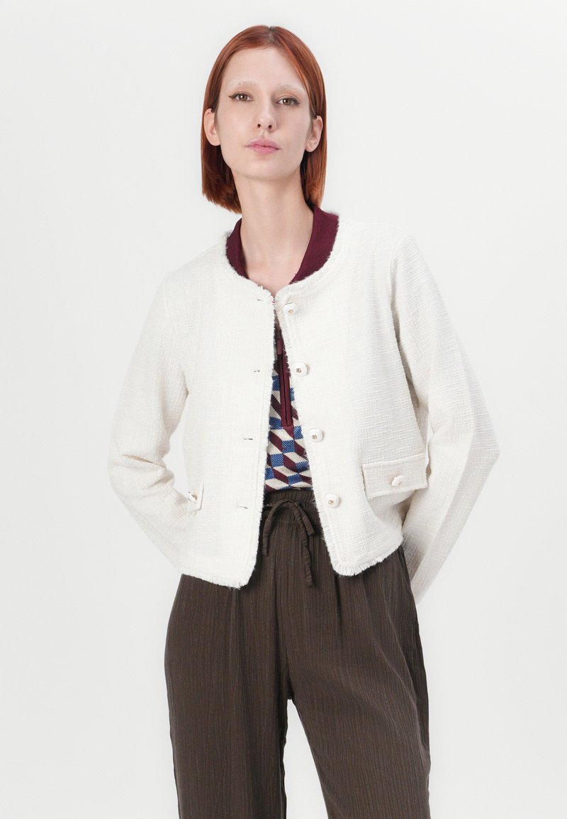 Witte textuurjacket met een ronde halslijn, voorzien van een bordeauxkleurige kraag, voor knopen en twee zakken. Gecombineerd met bruine geplooide broeken.