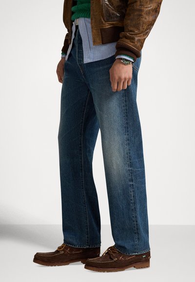 Μπλε τζιν παντελόνι με χαλαρή εφαρμογή, με ξεθωριασμένο πλύσιμο. Το μοντέλο φοράει καφέ μοκασίνια και στρωτά ρούχα, συμπεριλαμβανομένου ενός πράσινου πουλόβερ.