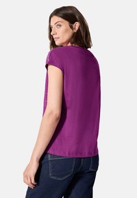 Top púrpura de manga corta con ajuste relajado, textura suave y escote redondeado. Combinado con unos jeans de mezclilla oscuros con costuras visibles.