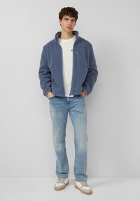 Młody mężczyzna w niebieskiej polarowej kurtce na zamek, białej koszulce, jasnoniebieskich dżinsach i białych trampkach z beżowymi podeszwami, stoi na tle gładkiej ściany.