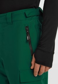 Green cargo pants with a black zippered pocket and a reinforced waistband. The fabric is smooth with a matte finish, featuring stitched accents.