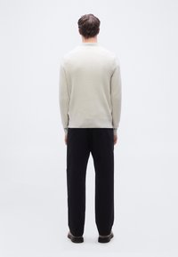 Homme debout de dos, portant un pull beige clair, un pantalon noir et des chaussures marron, sur un fond blanc uni.