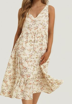 Ærmeløs kjole med V-hals i cremefarve med pink blomstermønster, lagdelt nederdel, båret af en person, der holder kjolens kant på den ene side.