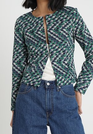 Vrouw draagt een cropped jas met een patroon in groen, blauw en wit, over een witte top met een hoge taille blauwe spijkerbroek.