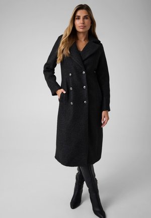 Une femme porte un long manteau noir croisé à boutons argentés et des bottes noires montant jusqu’aux genoux, sur un fond gris uni.