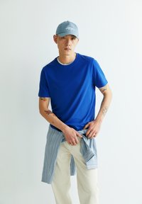 Blaues Rundhals-T-Shirt kombiniert mit hellfarbener Jeans, dazu ein gebundenes blau-weiß gestreiftes Hemd um die Taille. Die Kappe ist hellblau.