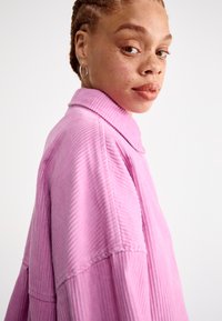 Hnedá košeľa z kordu v svetloružovej farbe s širokým golierom, textúrovanými vertikálnymi prúžkami a voľným strihom. Model pózuje v bočnom profile.