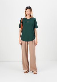 Kvinne står med ansiktet vendt fremover iført mørkegrønn GAP t-skjorte, beige vid bukser, brune sneakers og brun skulderveske mot hvit bakgrunn.