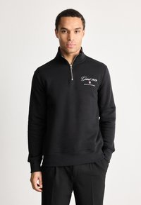 Muž v čiernom svetríku s krátkym zipsom a logom 'Gant 1949 American Sportswear' na hrudi, stojaci na jednofarebnom bielom pozadí.