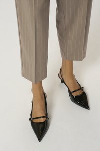 Svarta slingback-klackar i lackläder med spetsig tå och ankelrem, kombinerade med taupefärgade byxor med smala ränder.