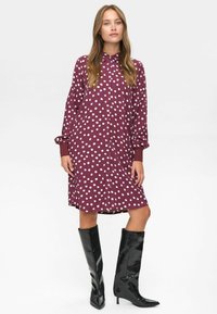 Femme portant une robe maroon à pois blancs et à manches longues, associée à des bottes en cuir noires jusqu'aux genoux.