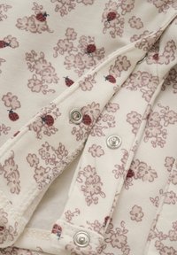 Tissu en coton avec un fond crème, présentant un motif floral répété avec des coccinelles rose pâle et rouge. Boutons-pression en argent présents.
