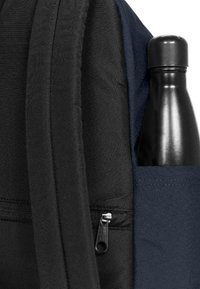 Sac à dos noir et marine avec une poche latérale contenant une bouteille d'eau noire, un tissu texturé et un détail de fermeture éclair visible.