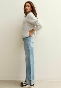 Jonge vrouw met losse krullend haar staat zijwaarts, gekleed in een lichtgrijze fuzzy trui, lichtblauwe opgerolde spijkerbroek en zwart-witte sneakers.