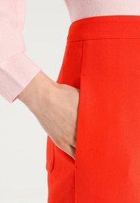 Rode stoffen rok met een gestructureerd oppervlak, voorzien van een zijzak. Gedempt roze langemouwen blouse zichtbaar aan de bovenkant.