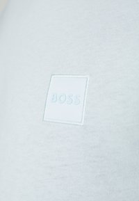 T-shirt en coton bleu clair avec un patch carré texturé portant le mot "BOSS" en lettres bleu clair, sur un fond de tissu lisse.