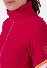 Fleecová bunda v jasně růžové barvě s vysokým límcem a plným zipem. Má oranžové a bílé pruhované akcenty na rukávech a odznak loga.
