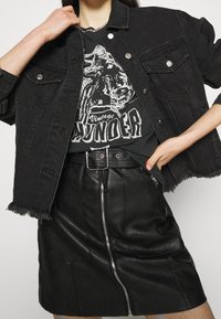 Czarna dżinsowa kurtka z postrzępionym dołem, zszyta z vintage'owym t-shirtem z grafiką. Czarna skórzana spódnica z przednim zamkiem błyskawicznym i akcentami w postaci paska.
