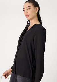 Črna bluzica z dolgimi rokavi in V-izrezom, gladka tekstura in stranske razpoke, v kombinaciji s temnimi jeans hlačami. Model drži torbico na tanko naramnico.