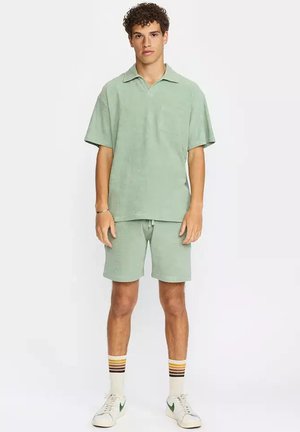 Junger Mann trägt ein mintgrünes kurzärmeliges Poloshirt und passende Shorts, weiße Sneaker und gestreifte Socken, steht vor einem weißen Hintergrund.