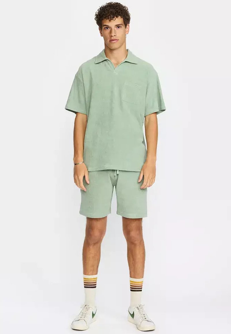 Joven con polo de manga corta verde menta y pantalones cortos a juego, zapatillas blancas y calcetines a rayas, de pie frente a un fondo blanco.