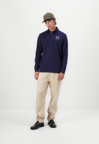 Polo de manga longa em azul-marinho com colarinho e logo 'H' bordado. Calças retas em bege e mocassins pretos, complementados com uma capa xadrez.