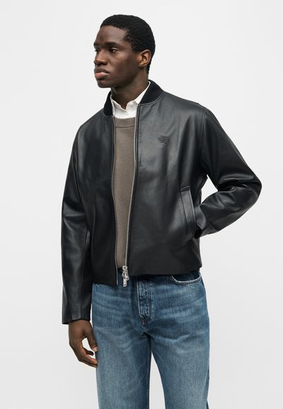 Άνδρας που φοράει μαύρο δερμάτινο μπουφάν τύπου bomber πάνω από μπεζ πουλόβερ και λευκό πουκάμισο με γιακά, συνδυασμένο με μπλε τζιν, κοιτάζοντας προς το πλάι.