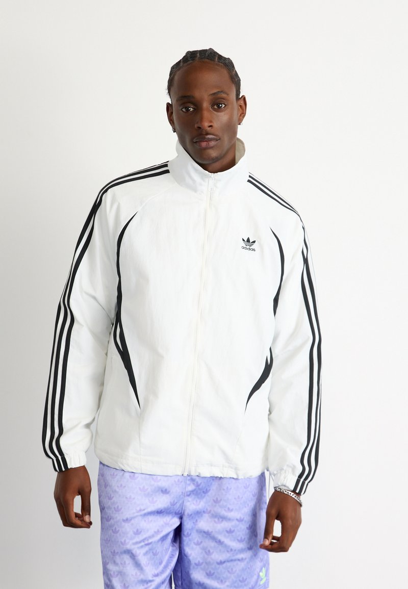 Bijela jakna na zip s crnim trakama duž ramena i ruku, s malim crnim Adidas logotipom na gornjem lijevom dijelu prsiju.