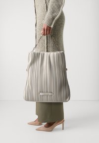 Gevouwen tote bag in off-white met verticale strepen, kettinghandvaten en een goudkleurige accent aan de basis. Zachte textuur, ruime uitstraling.