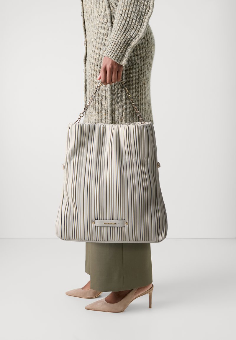 Gevouwen tote bag in off-white met verticale strepen, kettinghandvaten en een goudkleurige accent aan de basis. Zachte textuur, ruime uitstraling.