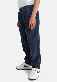 Pantaloni in corduroy blu navy con vita regolabile, gambe dritte e dettagli sulle cuciture laterali. Abbinati a sneaker bianche con accenti verdi.