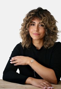 Silberarmband mit einem runden Charm, das ein Ausschnittdesign aufweist, getragen an einem Handgelenk mit langen, dunklen Ärmeln. Lockige Haare rahmen das Gesicht.