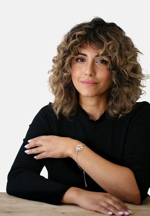 Bracelet en argent avec un pendentif circulaire arborant un design découpé, porté au poignet avec de longues manches sombres. Cheveux bouclés encadrant le visage.