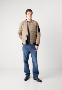 Jaqueta bomber acolchoada bege com um detalhe de bolso preto, usada sobre uma blusa azul-marinho, combinada com jeans azuis e sapatos pretos.