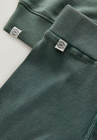 Zaļš ribots audums ar gludu tekstūru. Ar divām etiķetēm stūros, kas attēlo zīmola logotipu, uz piedurknēm un biksēm.