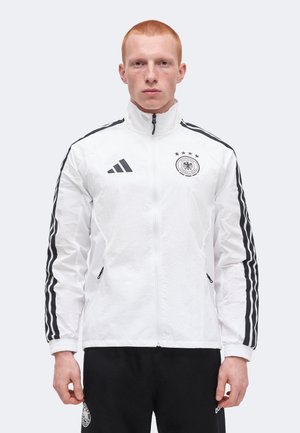 Homme aux cheveux rouges courts portant une veste Adidas blanche avec des bandes noires et l'emblème de l'équipe de football d'Allemagne, debout devant un fond uni.
