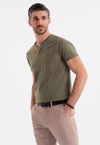 Camisa Henley verde de manga corta con bolsillo y tapeta de tres botones, combinada con pantalones de tono claro y un cinturón negro. Textura de algodón suave.