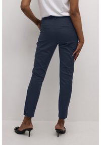 Pantalon bleu marine à coupe ajustée, avec des poches arrière et une texture de tissu lisse. Porté avec des sandales à talons noires.
