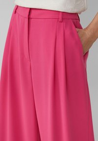 Pantaloni a vita alta con pieghe in tessuto rosa vivace. Presentano tasche laterali, una texture liscia e una vestibilità su misura per una silhouette slanciata.