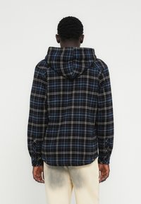 Καρό πουκάμισο φανέλας σε ναυτικό μπλε με γκρι, μπλε και λευκές αποχρώσεις, διαθέτει κουκούλα, μακριά μανίκια και κουμπωτά μανίκια, εμφανιζόμενο από πίσω.