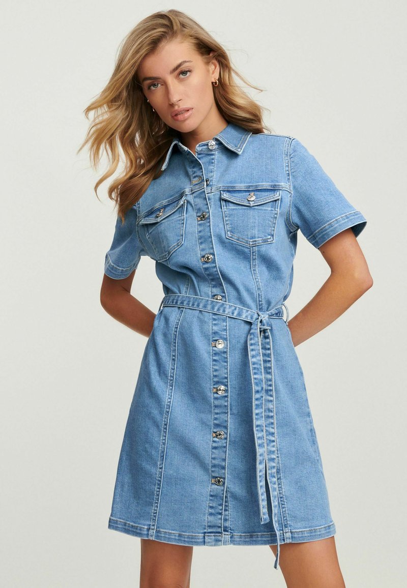 Das Denim-Shirtkleid verfügt über kurze Ärmel, eine durchgehende Knopfleiste, zwei Brusttaschen und einen abnehmbaren Bindegürtel, in einem hellblauen Farbton mit kontrastierenden Nähten.